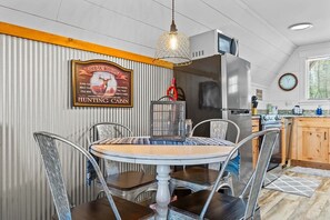 Un comedor con una mesa redonda, sillas metálicas, una lámpara colgante, una cocina al fondo y un letrero en la pared.
