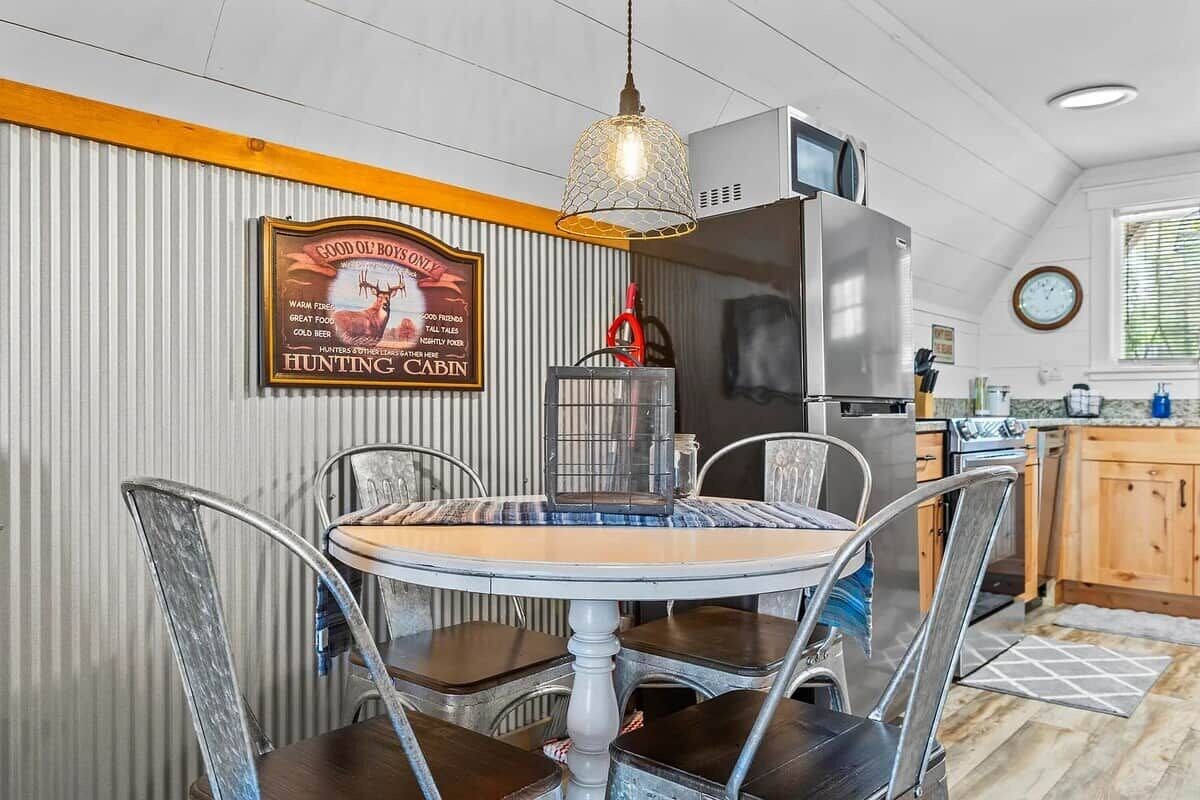 A dining area with a round table and metal chairs, a hanging light fixture, a refrigerator, and a kitchen counter with cabinets.