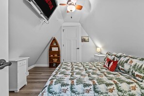 Un dormitorio con cama, televisor montado en la pared, ventilador de techo, estantería y una puerta.