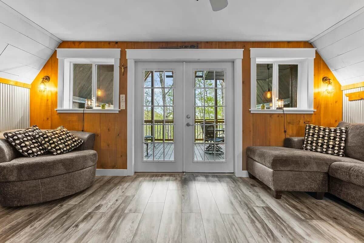 Quarto com decoração em madeira, dois sofás com almofadas estampadas, porta dupla com painéis de vidro e vista para o exterior.