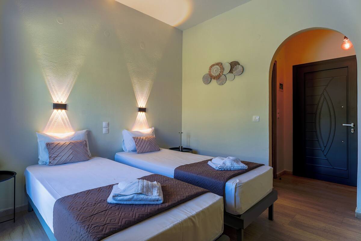 Habitación de hotel con dos camas, una mesita pequeña, una lámpara de pared y una puerta.