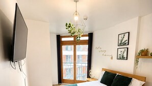Camera da letto moderna con un letto in legno, una TV a schermo piatto montata a parete, un'ampia finestra con vista sugli edifici e un lampadario.