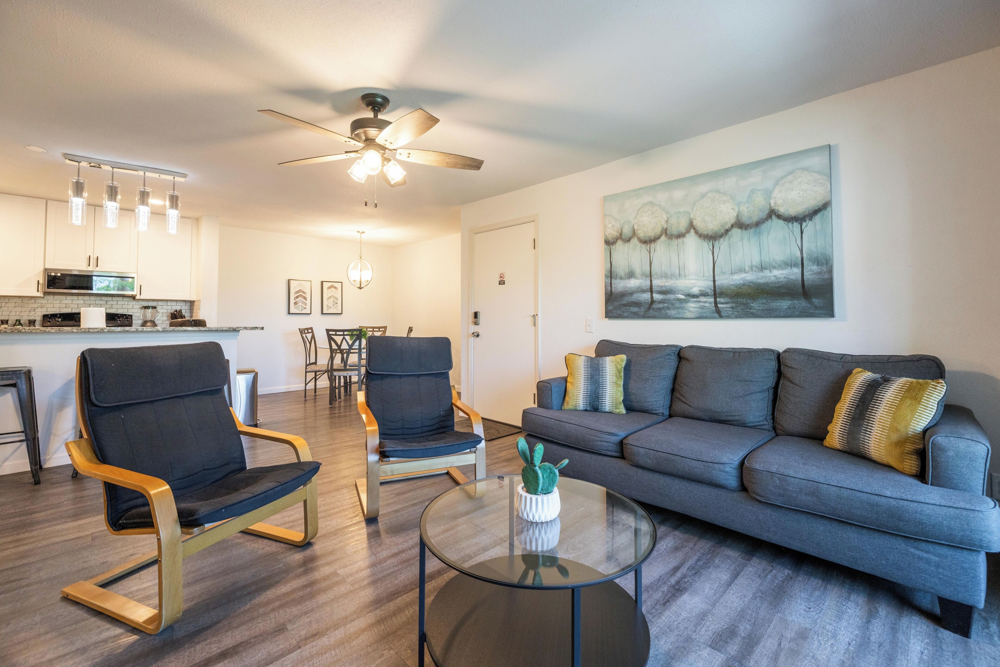 A modern living room with a gray sofa, two chairs, a glass coffee table, and a ceiling fan.