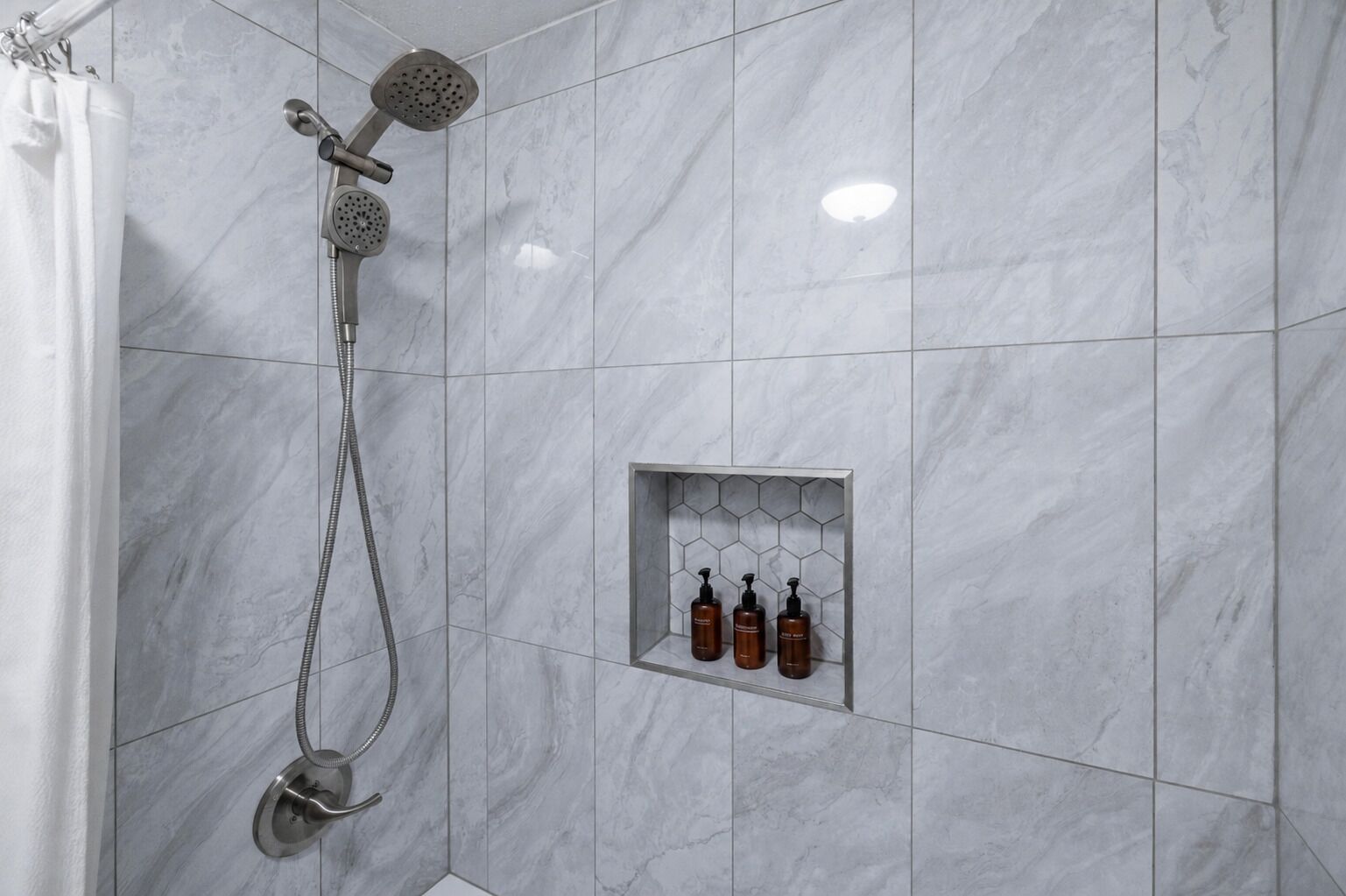 A shower with a handheld showerhead and a wall-mounted head, a shower curtain, and a shelf with three bottles of shampoo.