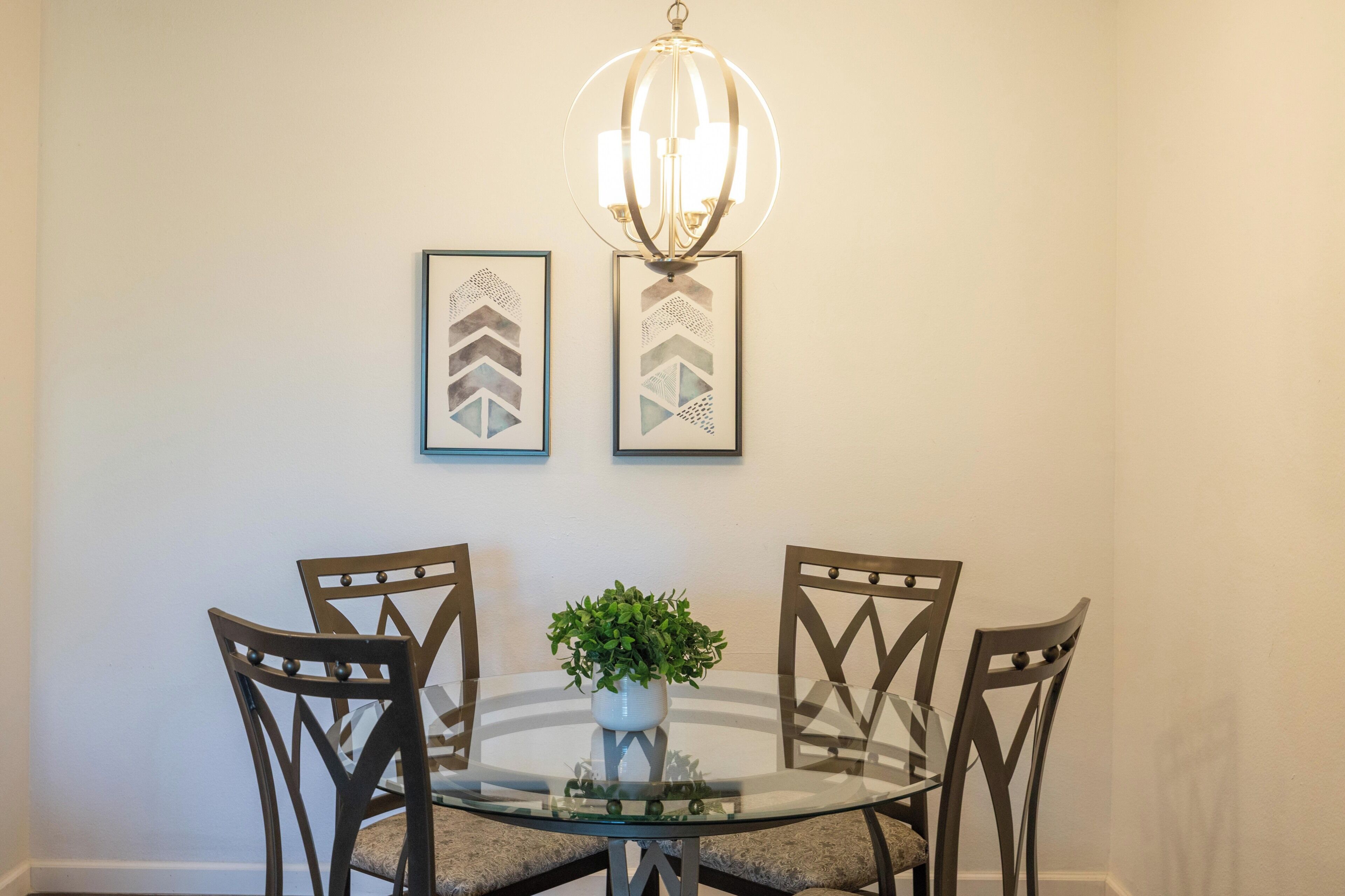 A dining area with a glass table, four chairs, a potted plant, and two framed artworks on the wall.