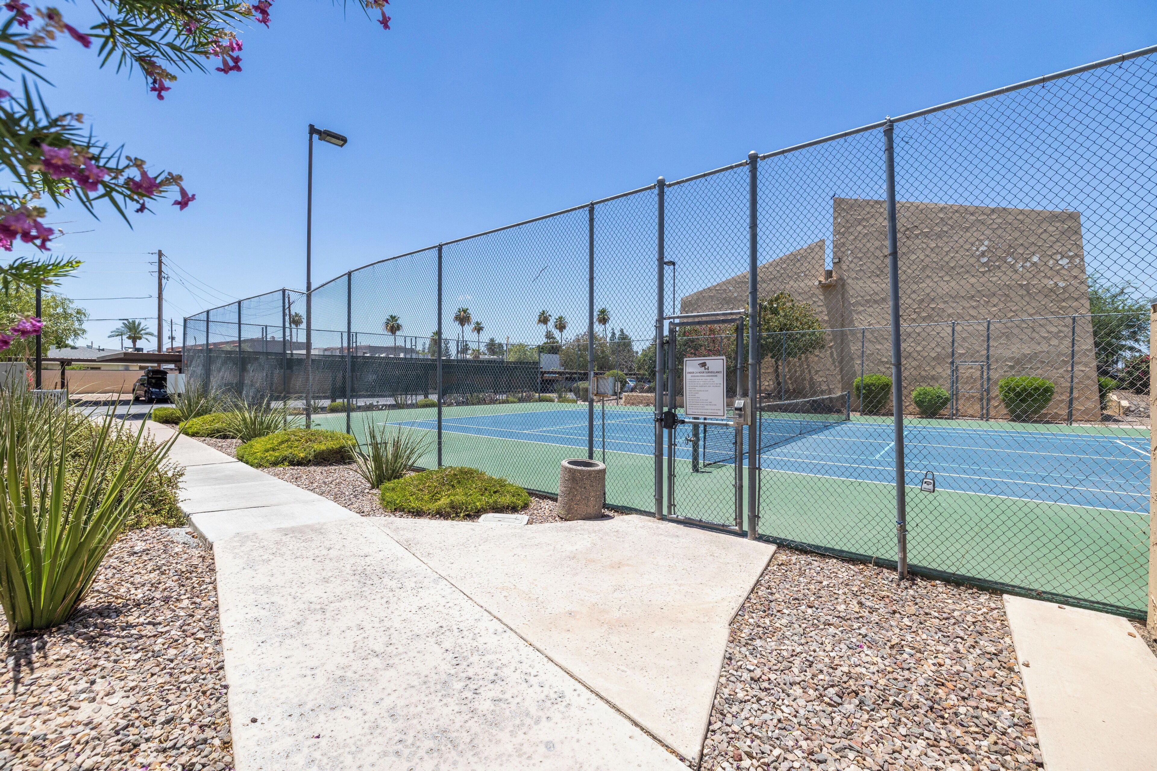 Un terrain de tennis entouré d’une clôture, avec un ciel dégagé, un chemin et un bâtiment en arrière-plan.