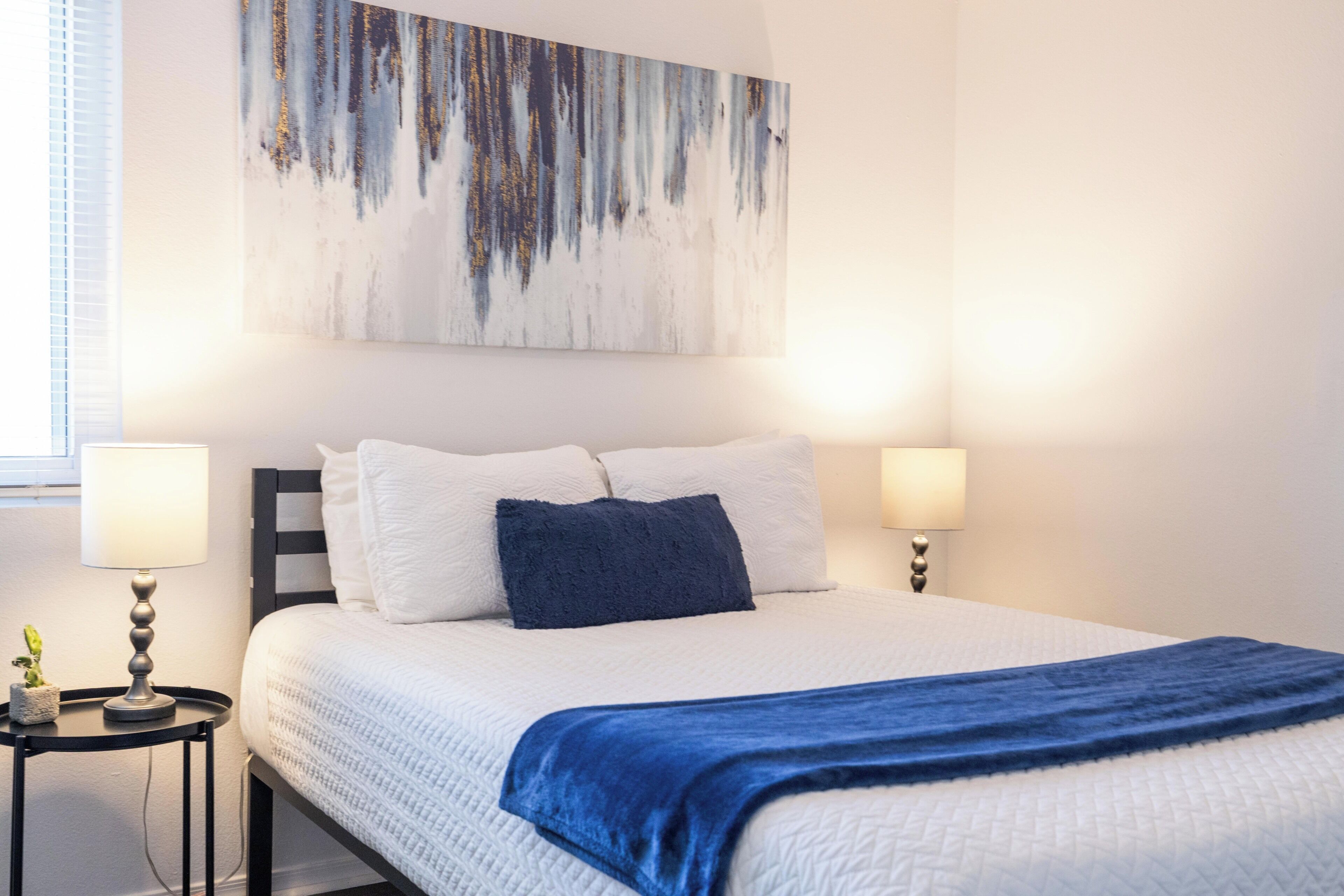 Een hotelkamer met een bed, twee bedlampen, een aan de muur hangend schilderij en een tafeltje met een plant.