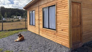 Eine Holzhütte mit zwei Fenstern und einer Tür, eine Schotterauffahrt und ein Hund, der draußen liegt.