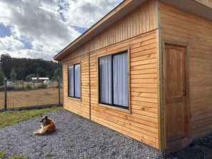 A wooden cabin with two windows and a door, a gravel driveway, and a dog lying outside. - Spacious 3D family cabin plus ample parking, beautiful nature. (Villarrica)