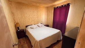 Ein Zimmer mit Holzwandverkleidung, einem Bett, einem Nachttisch, einer Lampe und einem violetten Vorhang.