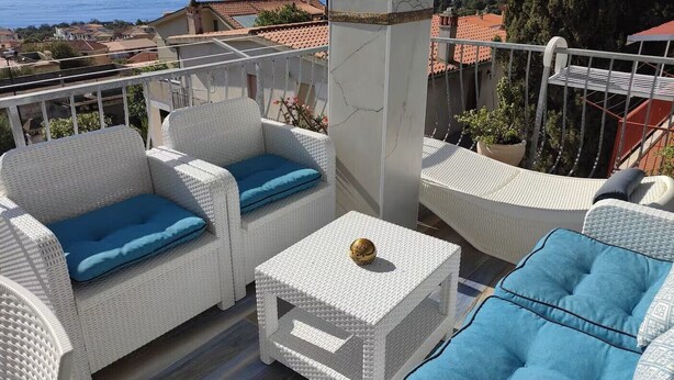 A balcony with wicker furniture, a marble column, and a view of the sea and buildings. - Dalhia's House: stunning gulf and mountain views, Wi-Fi, A/C Free parking (Cala Gonone)
