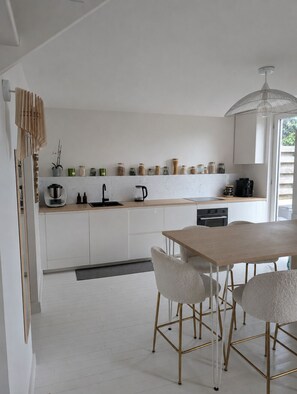 A modern kitchen with white cabinets, a wooden countertop, and a dining area with a table and chairs. - Charming house 500 m from the beach - Kerroc'h (Ploemeur)