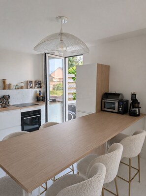 Una cocina moderna con una larga mesa de madera, sillas blancas, horno empotrado y cafetera.