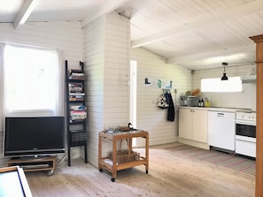 Un espace de vie compact comprenant un téléviseur, une bibliothèque, un chariot en bois, un coin cuisine et une fenêtre.