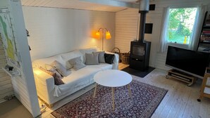 A cozy living room with a white sofa, patterned cushions, a small round table, a TV, a fireplace, and a window with a view of greenery. - Charming 2-bedroom cottage in Kulhuse, with a wonderful garden, close the beach (Kulhuse)