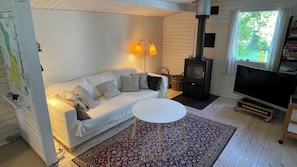 A cozy living room with a white sofa, patterned cushions, a small round table, a TV, a fireplace, and a window with a view of greenery.