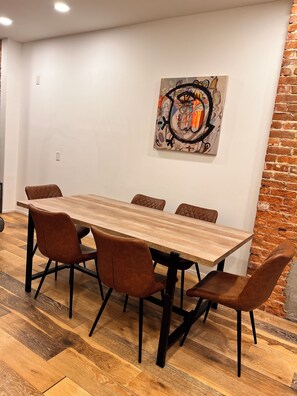 A dining area with a wooden table and six brown chairs, a brick wall, and a colorful abstract painting on the wall. - 5BR Jersey City Home – Perfect for World Cup Groups! 
Walk to the PATH train. (Jersey City)