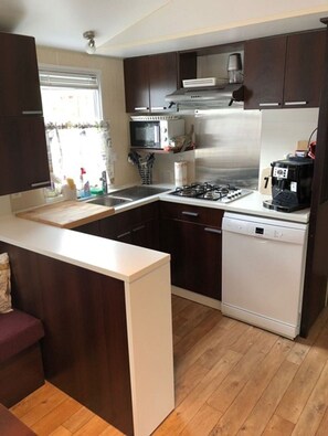 A compact kitchen with dark wood cabinets, a stainless steel range hood, a microwave, a gas stove, and a dishwasher. - Mobile home for rent at the Plage de Pampelonne St. Tropez-Ramatuelle (Saint-Tropez)