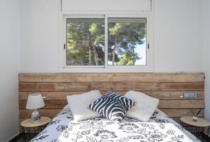 A bedroom with a wooden headboard, a window with a view of trees, a bed with floral bedding, a bedside lamp, and a small table with a vase.