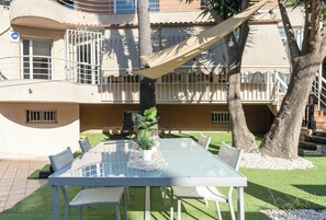 Mesa con cubierta de vidrio, sillas blancas, una planta en maceta y una hamaca en un patio exterior con un edificio al fondo.