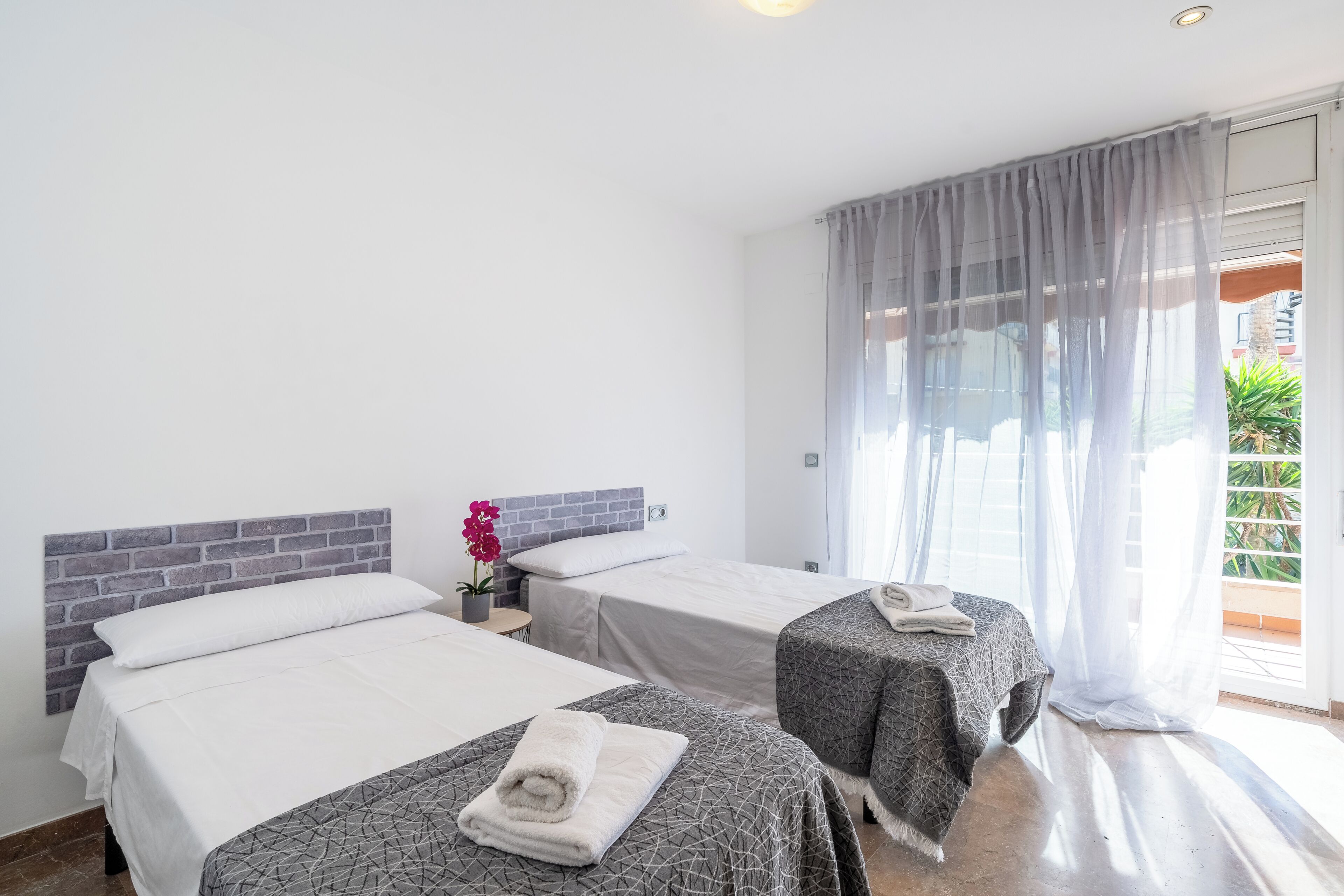 Ein Hotelzimmer mit zwei Betten, einem Ziegelmuster-Kopfschild, einer Topfpflanze und Blick durch transparente Vorhänge.