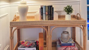 Ein Zimmer mit einem runden Spiegel, einer Lampe, einem Korbstuhl-Konsolentisch mit Büchern und Dekoration sowie einer kleinen Pflanze.