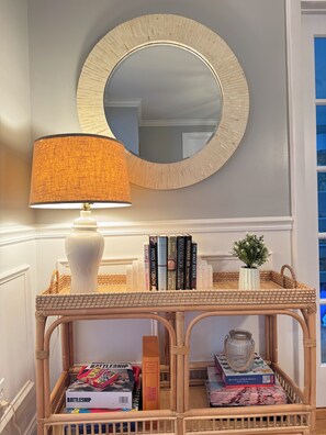 A room with a round mirror, a lamp, a wicker console table with books and decorative items, and a small plant. - Central Wakefield Home with Spacious Outdoor Living and AC (South Kingstown)