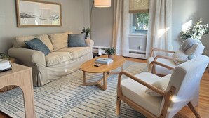Ein Wohnzimmer mit einer Couch, einem Couchtisch und zwei Sesseln, einem Gemälde an der Wand und einem Fenster mit Vorhängen.