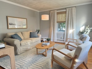 Ein Wohnzimmer mit einer Couch, einem Couchtisch und zwei Sesseln, einem Gemälde an der Wand und einem Fenster mit Vorhängen.