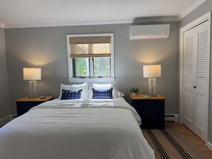 Quarto com cama, duas mesas de cabeceira, janela com persianas e ar condicionado, além de uma porta com venezianas.