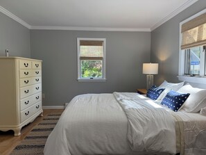 Ein Schlafzimmer mit Bett, Kommode, Fenster und Lampe, mit Blick auf Bäume.