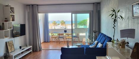 A living room with a sofa, a television, a bookshelf, a dining area with chairs, and a view of the sea through large windows.