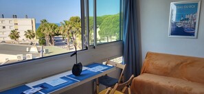 Ein Zimmer mit großem Fenster, Schreibtisch mit Papieren, Stuhl und einer Couch.