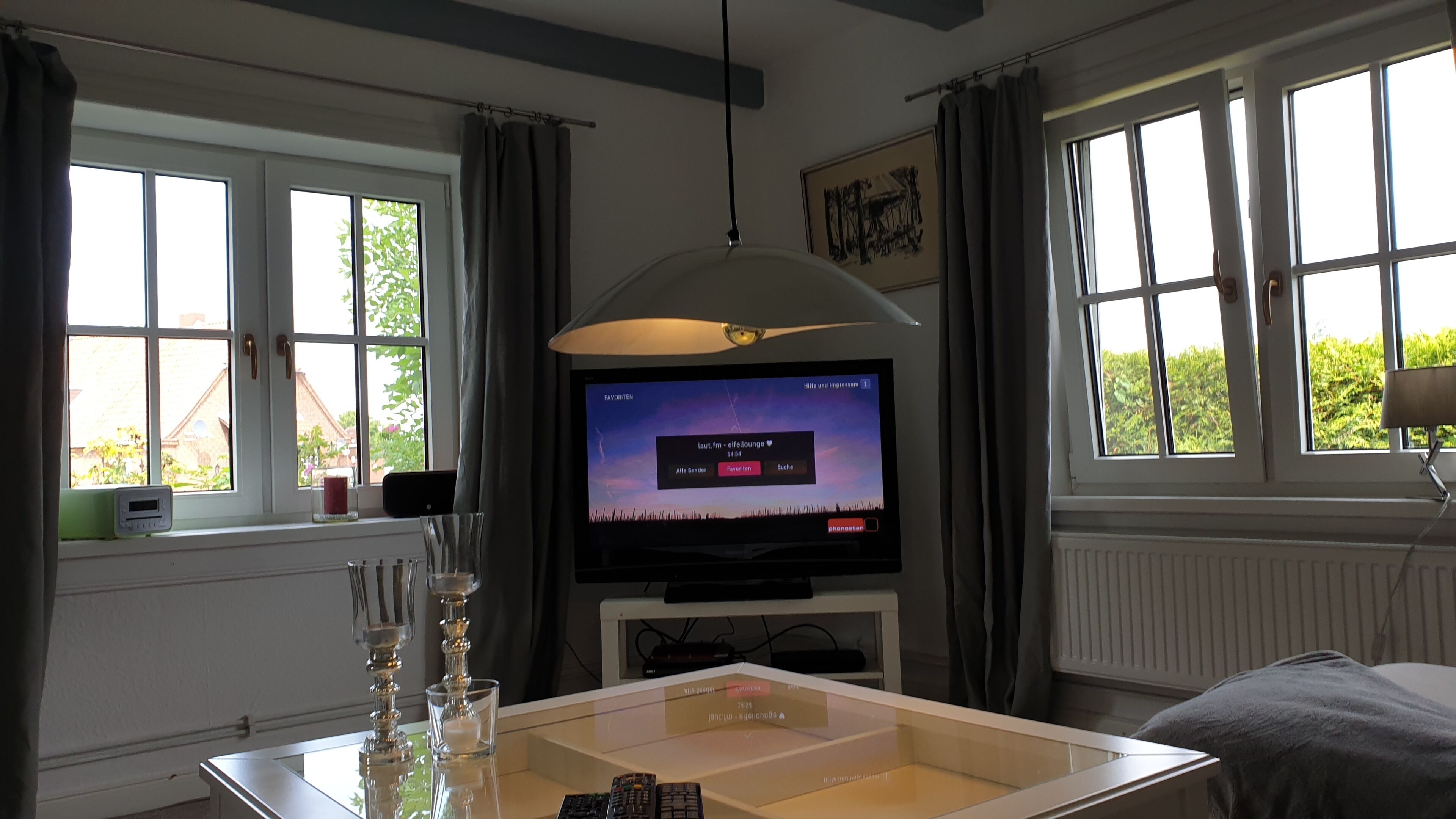 Ein Zimmer mit Fernseher, der einen Musikkanal zeigt, einem Glastisch mit Fernbedienung und einem Fenster mit Vorhängen.