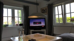 A room with a television displaying a music station, a glass table with a remote, and a window with curtains.