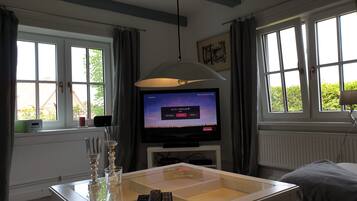 Ein Zimmer mit Fernseher, der einen Musikkanal zeigt, einem Glastisch mit Fernbedienung und einem Fenster mit Vorhängen.
