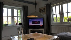 A room with a television displaying a music station, a glass table with a remote, and a window with curtains.