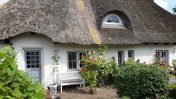 Ein Haus mit einem Strohdach, weißen Wänden, einer Bank und blühenden Pflanzen.