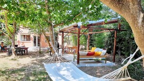 Una hamaca bajo una pérgola de madera, un banco de madera con cojines y una mesa de comedor con sillas en un jardín.