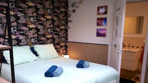 A neatly made bed with two rolled towels, a wall with a fish pattern, and a door leading to a bathroom.