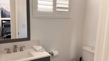 Un baño con lavamanos blanco, muebles oscuros, inodoro y ventana con persianas.
