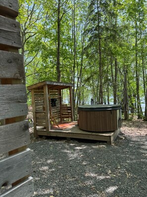 Una baita di legno con vasca idromassaggio e terrazza, immersa tra alberi e un lago.
