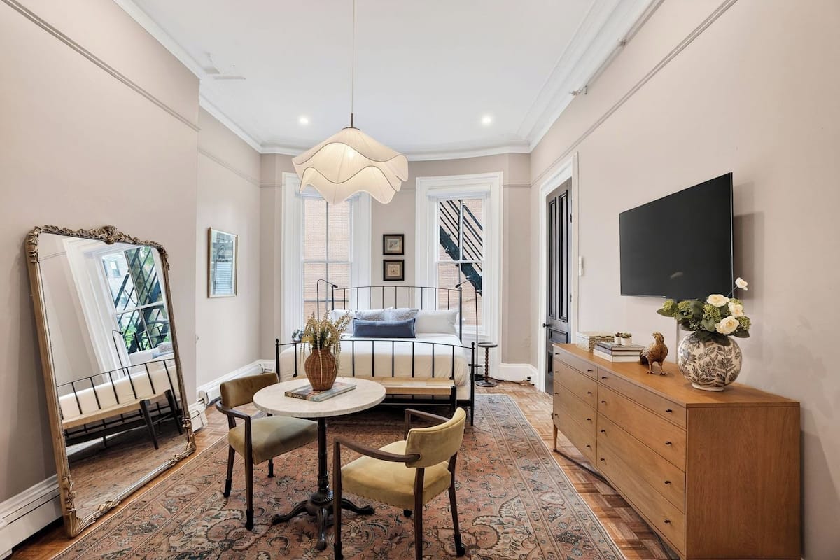 A room with a bed, a round table with chairs, a dresser, a mirror, and a TV on the wall.