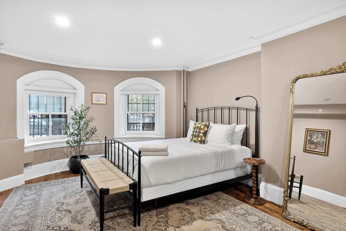 A bedroom with a bed, two windows, a potted plant, a bench, a floor lamp, and a mirror.