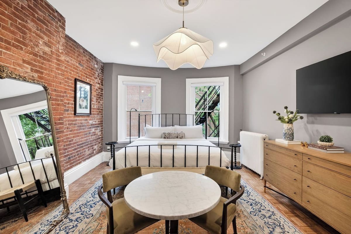 A bedroom with a bed, a round table, chairs, a dresser, a TV, and windows with a view of greenery.
