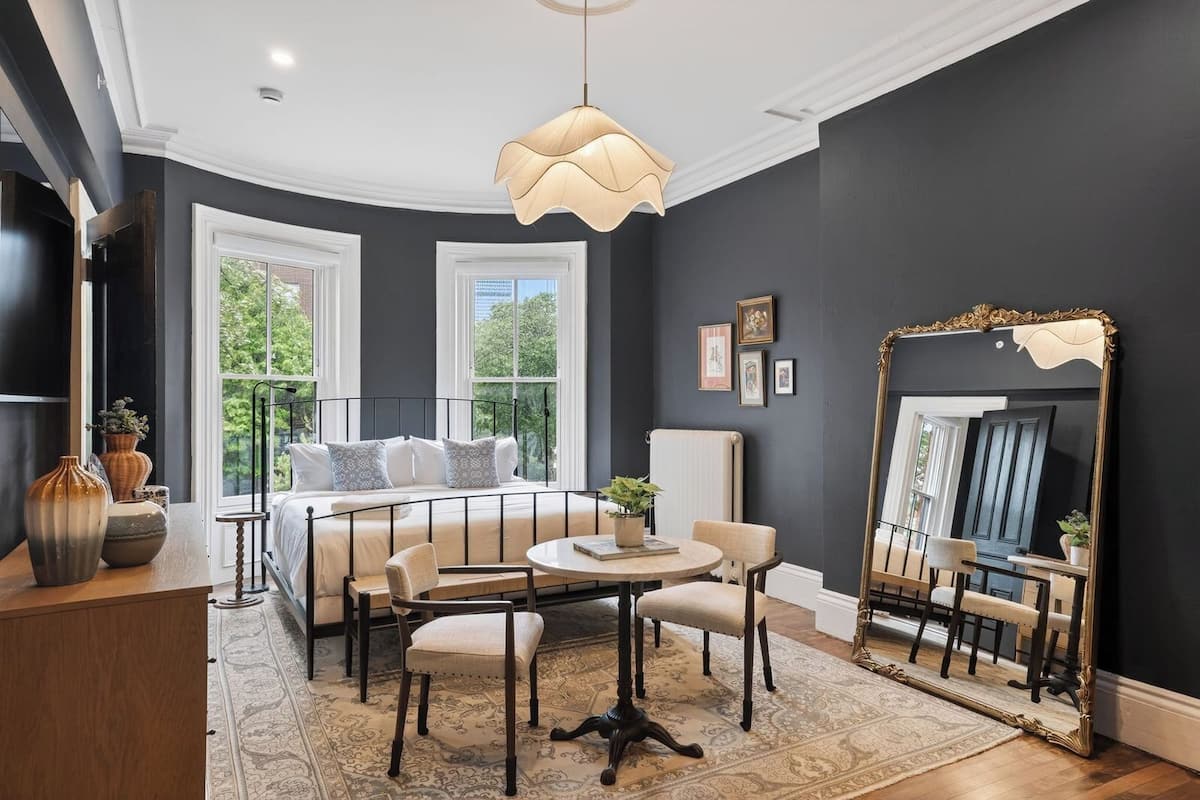 A room with a bed, a round table, chairs, a large mirror, and a view of greenery outside.