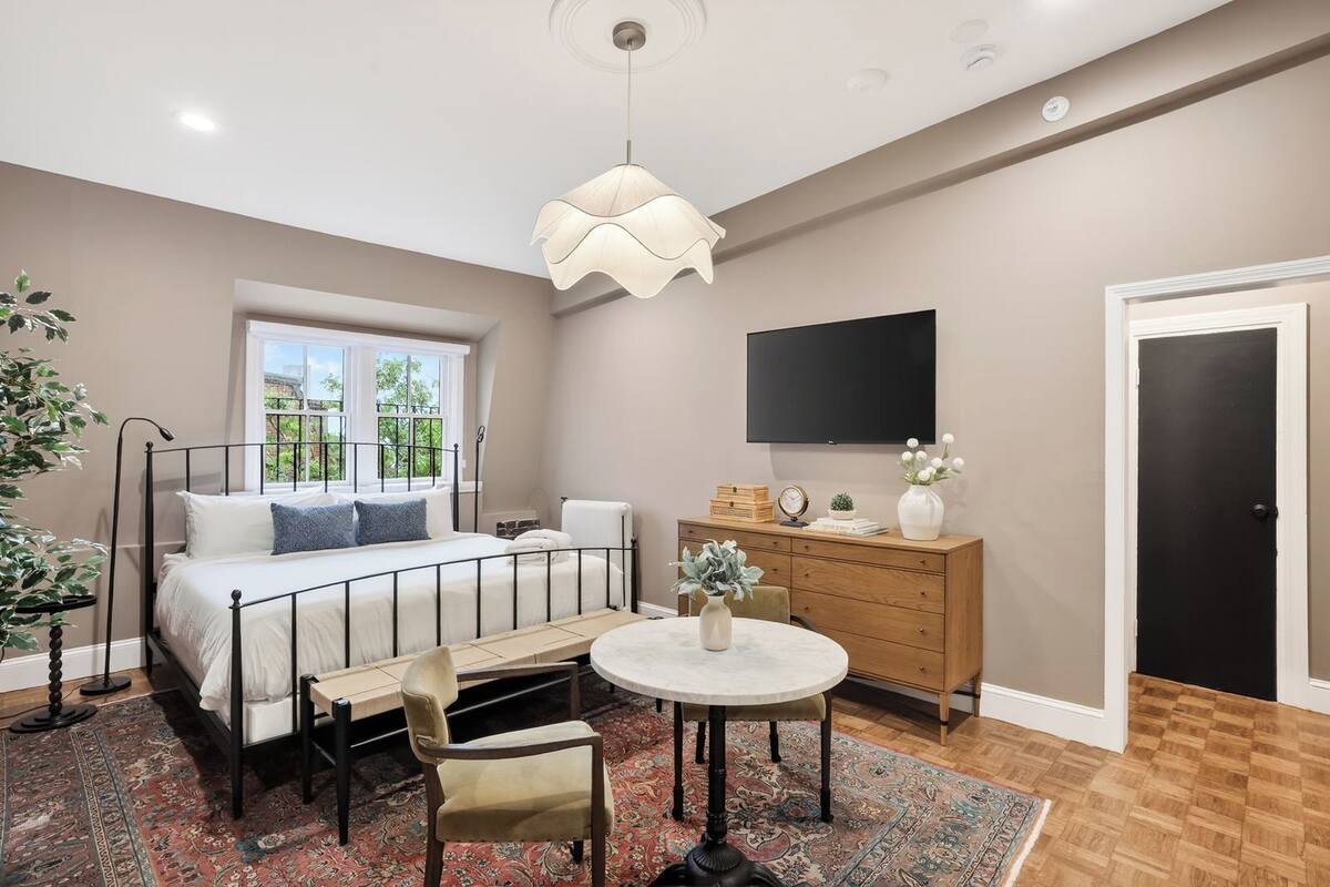 A bedroom with a bed, bedside table, chair, round table, dresser, TV, and a door.
