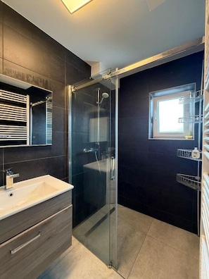 Een moderne badkamer met een douchecabine van glas, een witte wastafel, donkere tegels en een raam.