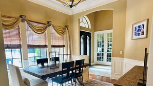 Uma sala de jantar com um candelabro, uma mesa, cadeiras, um tapete e vista para o exterior.