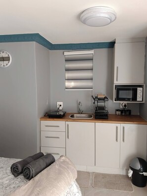 A compact kitchenette with a sink, microwave, and coffee maker, a clock, and a bed with rolled towels. - Crompton Studio Apartment 786 (Durban)